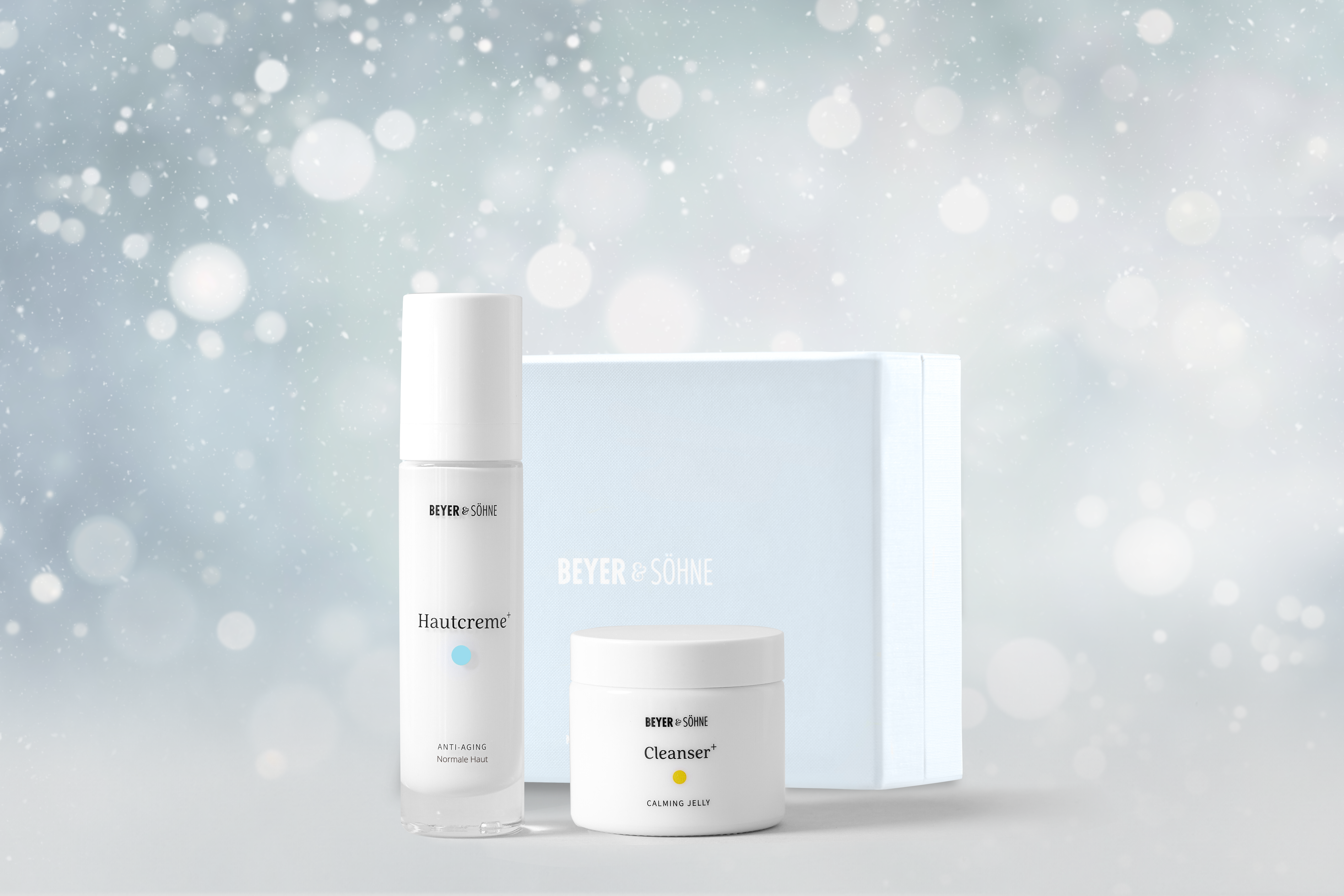 Hautcreme+ für normale Haut und Cleanser+ von Beyer & Söhne in hochwertigem Glasspender und Tiegel vor hellblauer Geschenkbox und weihnachtlich gestimmten Hintergrund mit Schneeflocken.